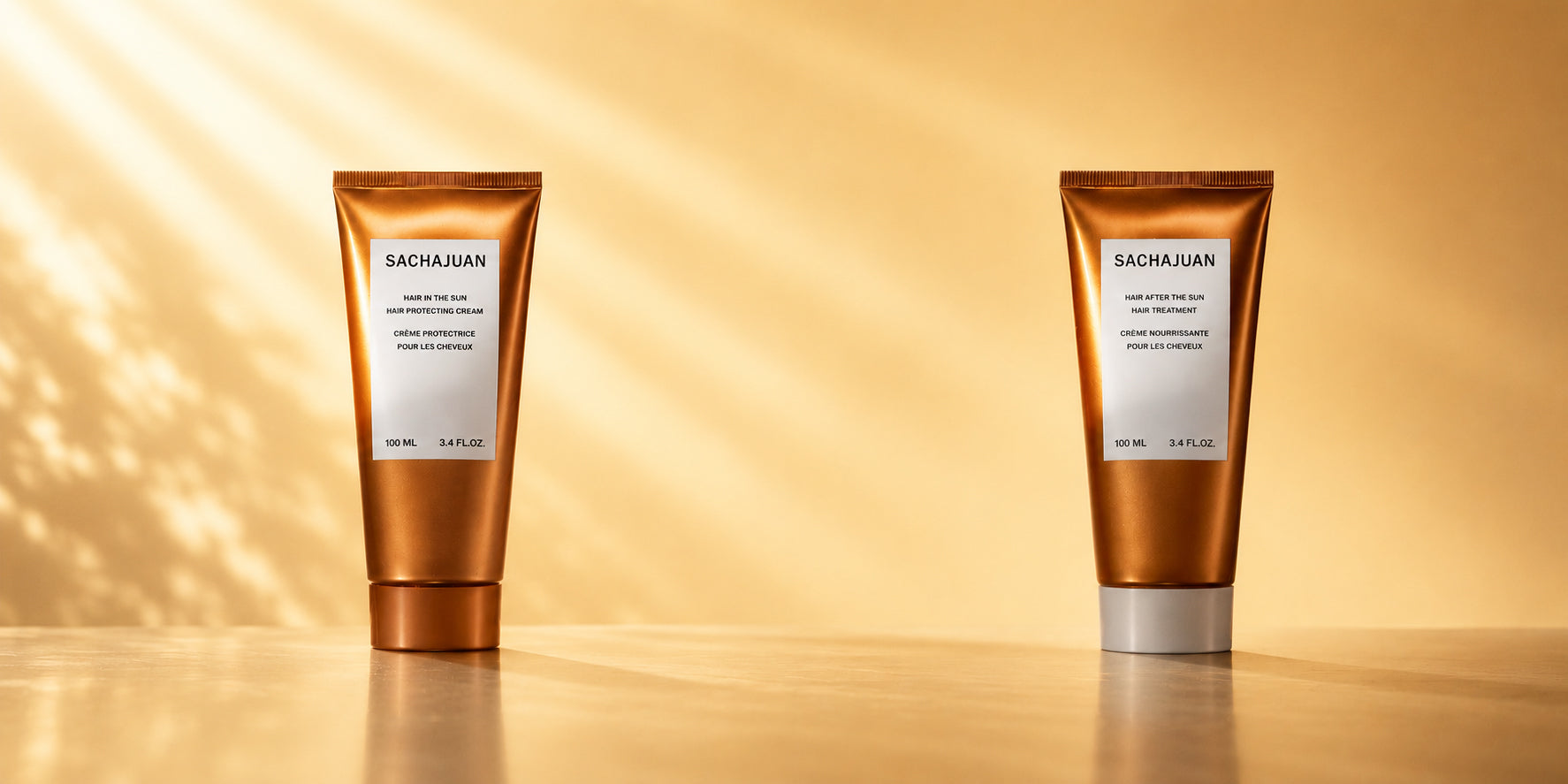 Two Sachajuan hair care tubes standing upright on a reflective surface against a warm golden background with soft light streaks.