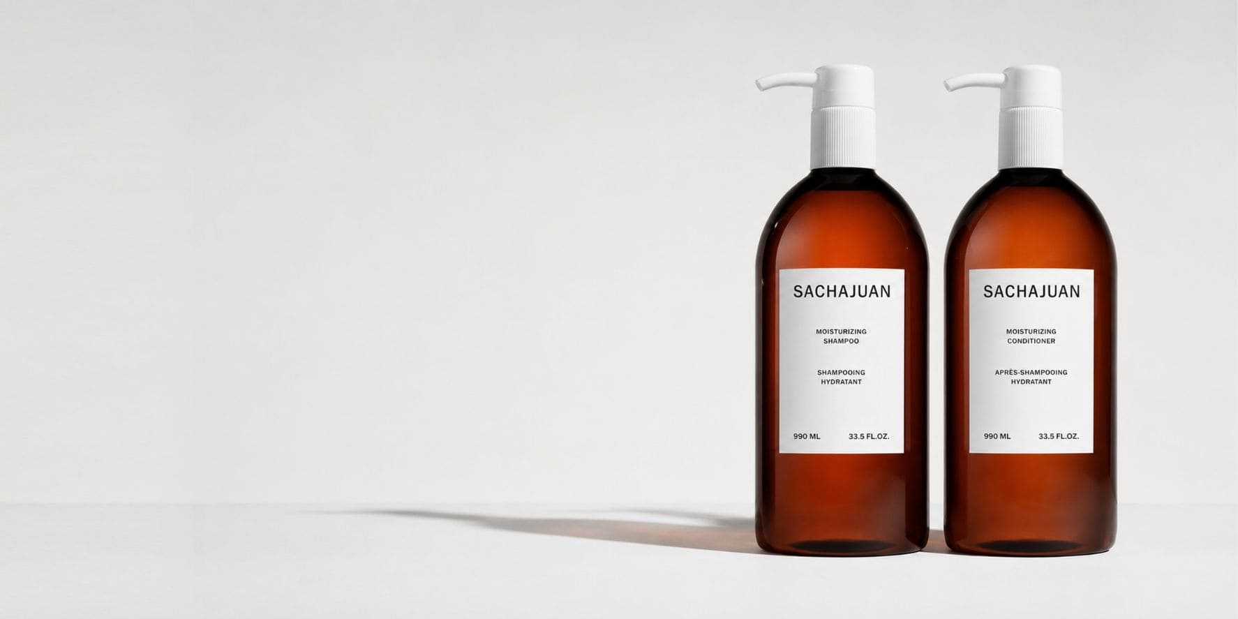 Two amber plastic pump bottles of Sachajuan haircare products with white labels, standing side-by-side on a light gray background.